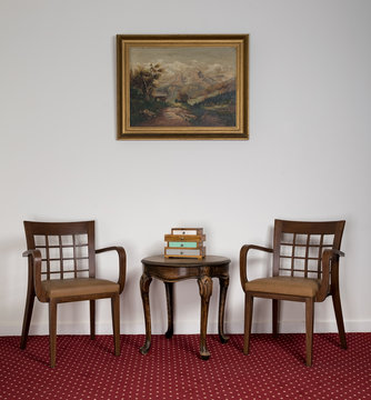 Composition of two wooden armchairs, small round coffee table and framed painting on red carpet and white wall