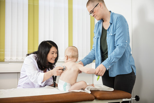 Mother Looking At Smiling Pediatrician Examining Baby On Bed