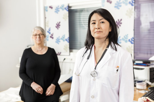 Confident Doctor Standing While Patient Sitting In Background