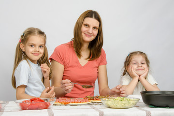 Mum with two little girls sitting at the kitchen table preparing a pizza