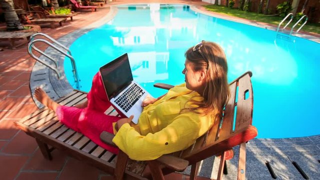 Blond Girl Sits on Folding Chair Works on Laptop by Pool