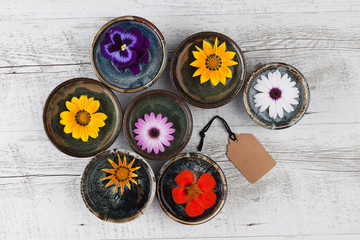 Colorful flowers floating in water in small ceramic bowls on rustic wooden table with gift tag. Top view with copy space.
