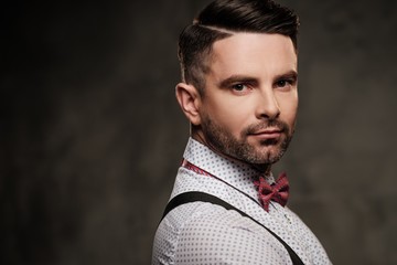 Stylish man with bow tie wearing suspenders and posing on dark background.