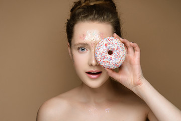 Portrait of young beautiful girl with donut in hand on the gold background. Nude makeup with tinsel. Concept of the beauty.