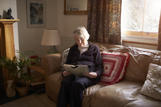 Senior Woman Sitting On Sofa Reading Book At Home