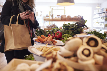 Woman Looks At Shopping List On Mobile Phone In Delicatessen