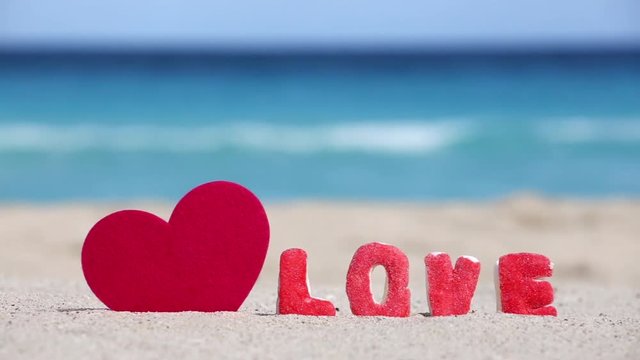 One pink heart shape and sign LOVE made of sweet letters on sandy tropical beach 
