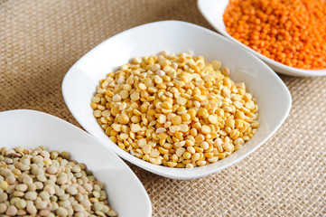 Colorful variety of cereals in a bowls on rustic burlap background. Red lentils, yellow lentils and green dried lentils 