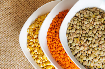Colorful variety of cereals in a bowls on rustic burlap background. Red lentils, yellow lentils and green dried lentils 