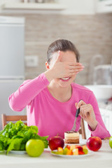 Young woman covering her eyes while eating a cake instead of healthy fresh fruit and vegetable on the table