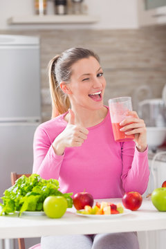 Happy young woman showing OK sign and winking while drinking fresh healthy smoothie in her kitchen