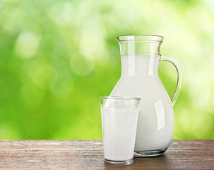 Pitcher and glass of milk on wooden table against blurred green background