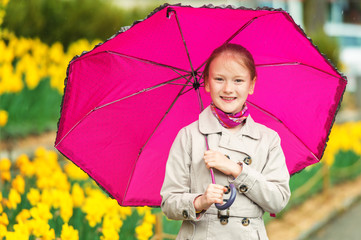 Outdoor portrait of adorable little girl of 7-8 years old, wearing beige trench coat, holding big pink umbrella, standing under the rain in a beautiful park with yellow narcissus