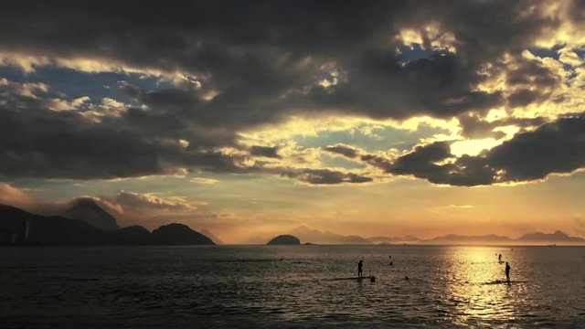 Scenic sunrise scene from Copacabana Beach featuring silhouettes of paddle boarders standing in front of the dramatic silhouette of Sugarloaf Mountain