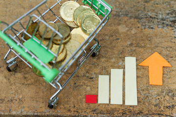 Trolley filled with coins beside chart showing increasing trend.. Concept of growth, consumerism,increasing trend,inflation,purchasing and spending power,economy.
