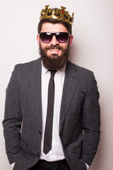 Young handsome man in sunglasses wearing suit and crown keeping hand on his jacket  while standing...