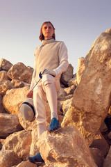 Portrait of a fencer man standing on top of the rock holding fencing mask and a sword on blue sky...