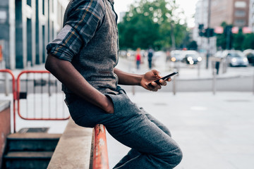 From the neck down view of young afro black man holding a smart phone, tapping the scree outdoor in...