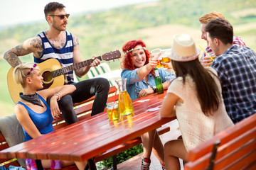 Girls toast with glasses of beer while tattooed boy play guitar