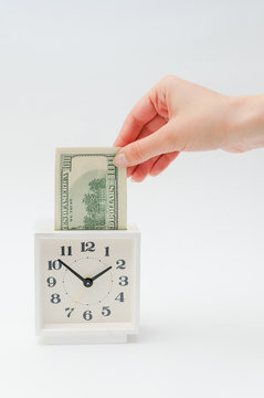 The person invests money in time. Investment business idea concept. Insertion of finance in time interval. White alarm clock moneybox. The female hand puts the note of hundred dollars an hours.