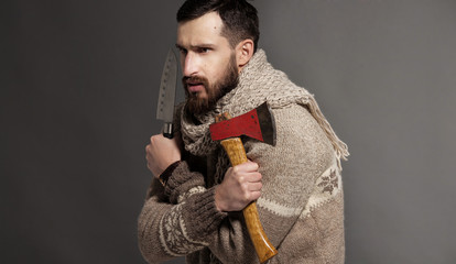 Evil young adult beaded man holding knife and axe looking at the camera. Fashion shot on grey background.