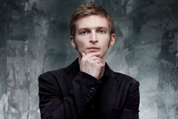 young man in jacket thoughtfully looks into the camera, hand on chin