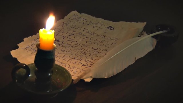 Ancient Manuscript and Burning Candle. The manuscript was written with a quill pen in an unknown alphabet. Next to the candle holder lit candle. 
