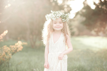 Smiling kid girl 4-5 year old wearing stylish white dress and flower wreath outdoors. Looking at...