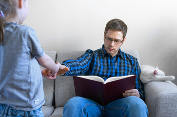 Little girl disturbing her dad while he is reading book.