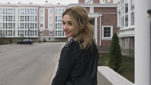 Young woman walking on the street. Blonde turns flirting gaze with the viewer. Black leather jacket