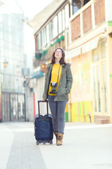 Smiling young hipster woman walking down the street of old town with a suitcase and vintage camera