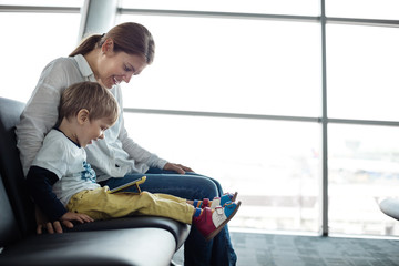 Little boy and his mother sitting in an airport departure hall and playing on a tablet