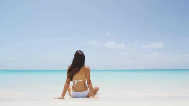 Vacation travel bikini woman on Caribbean beach. Young Lady with slim sexy body sitting on tropical white sand beach in Caribbean looking at perfect turquoise ocean. Luxury destination. SLOW MOTION.