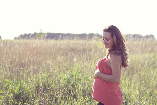 Young beautiful pregnant woman holding tummy smiling, in red a light summer dress, happy on meadow the grass 