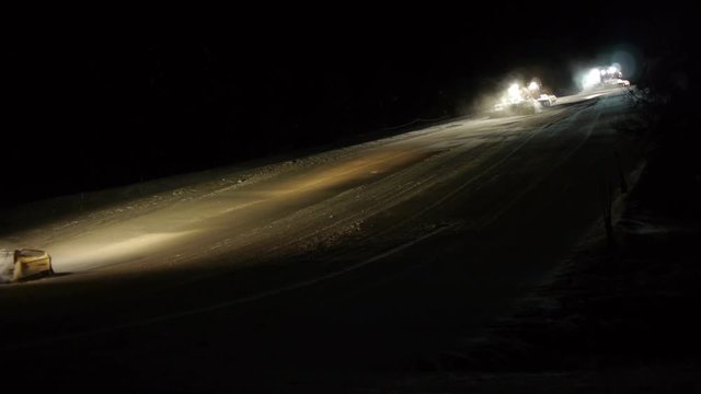 A couple of snow cats in line grooming snow up the ski slope at night time.
