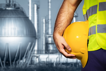 Hand or arm of engineer hold yellow plastic helmet in front of oil refinery industry
