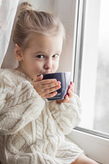 A little girl in the white knitted sweater sits on a window and drinks from a cup - 108538009