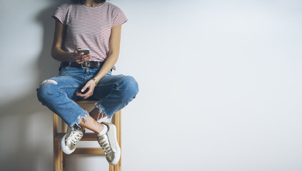 Close-up of young hipster girl wearing blue ripped jeans and using modern smart phone on a blank wall background with copy space for your text message or content