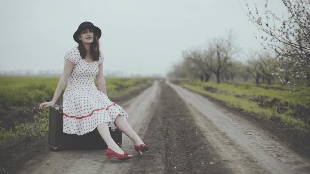 young woman dressed in retro style sitting on suitcase on the country road 