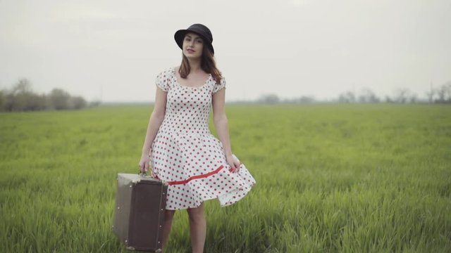 woman dressed in retro style poses with a suitcase on green field slow motion