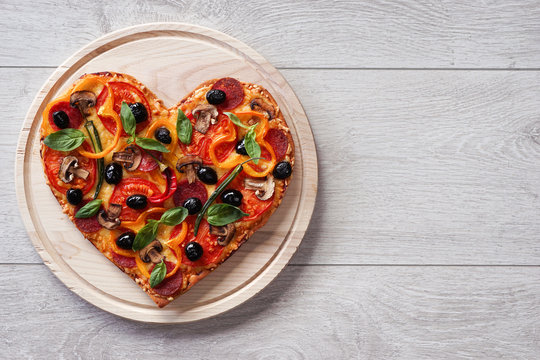 Baked heart-shaped homemade pizza on a cutting board on white wooden background. Close-up