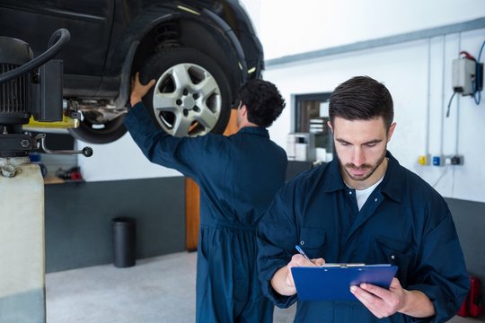 Mechanic preparing a check list while a colleague fixing cars wh