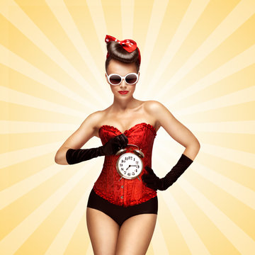 Time to sleep / Glamorous pinup girl in a red vintage corset holding a retro alarm clock in her hand and posing on colorful abstract cartoon style background.