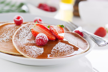 Homemade buttermilk american pancakes with fresh strawberry and raspberry on a white plate on a white table for breakfast, closeup, selective focus..