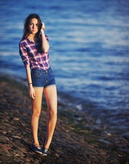 beautiful brunette girl in shorts and a plaid shirt standing on background of river embankment