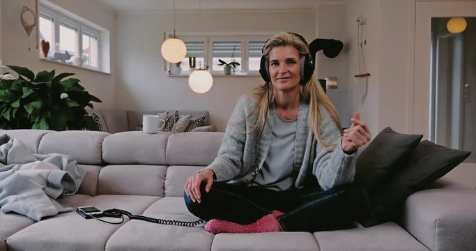Smiling happy woman relaxing with her music at home sitting cross-legged on a sofa listening to tunes off her mobile phone on a pair of stereo headphones
