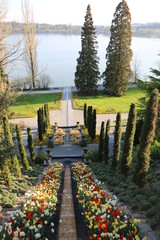 Kunstvolle Parkanlage im Fr&uuml;hling, im Hintergrund ein See, im Vordergrund ein mit Fr&uuml;hlingsblumen bepflanzter Wasserlauf, der im See m&uuml;ndet