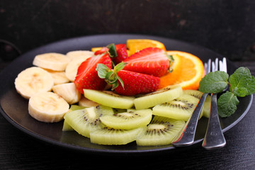 sliced fruit (strawberries, kiwi, orange, banana) on a black plate