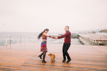 Young happy couple walking with dog and having fun on the rainy berth in autumn. Sea background