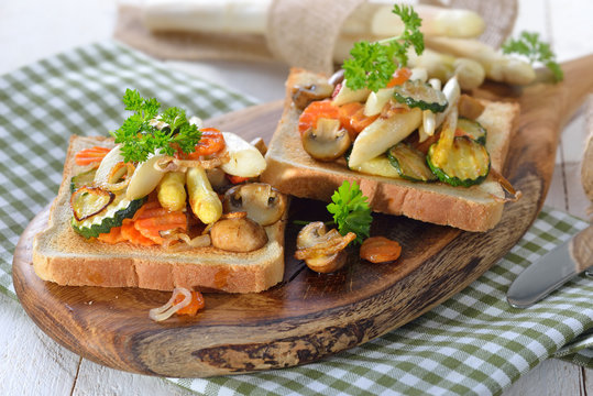 Vegetarischer Toast mit gebratenem weißen Spargel und gemischtem Gemüse, auf einem Holzbrett serviert - Vegetarian toast with fried white asparagus and mixed vegetables, served on a wooden board